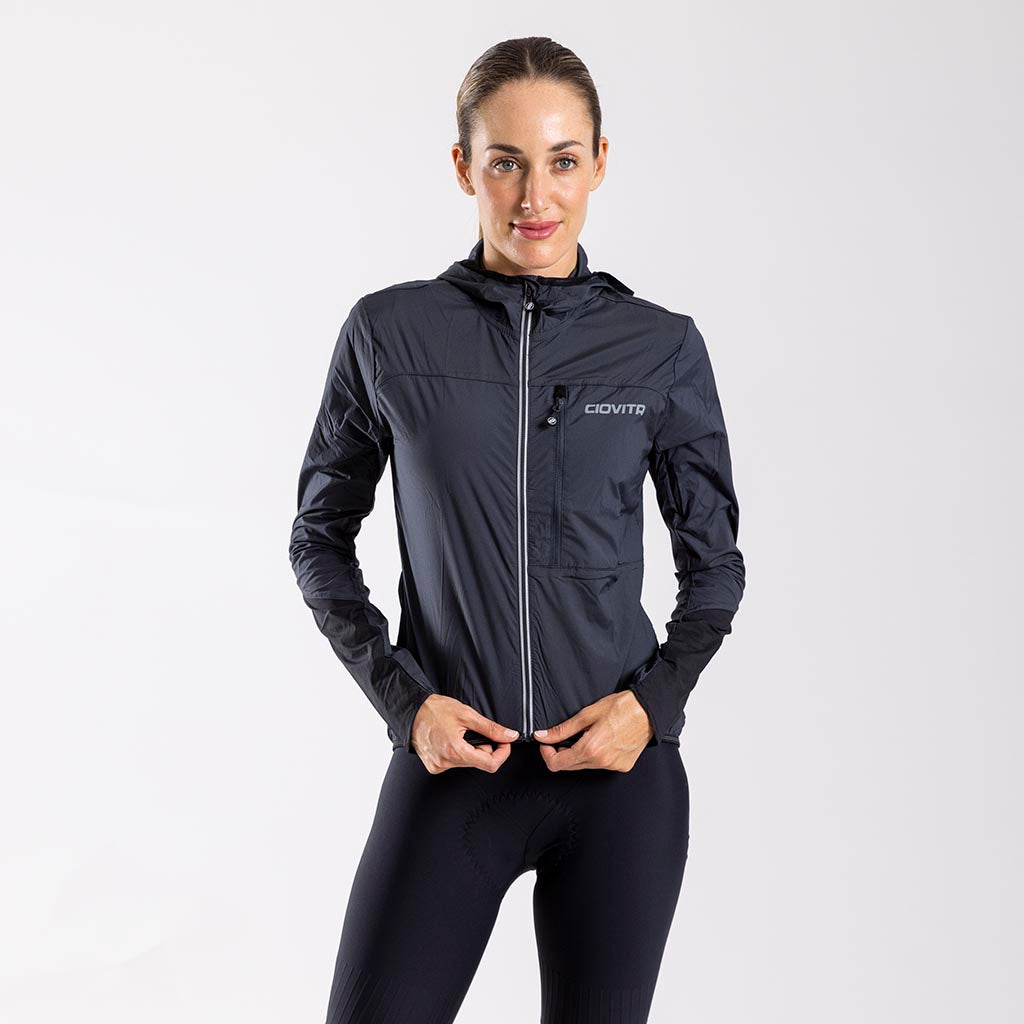 A woman wearing a charcoal cycling jacket with full length YKK zipper, designed for gravel road riding. The jacket features a windproof fabric, under helmet hood, reflective trims, and an elasticated hem.