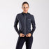 A woman wearing a charcoal cycling jacket with full length YKK zipper, designed for gravel road riding. The jacket features a windproof fabric, under helmet hood, reflective trims, and an elasticated hem.